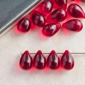 May include: A collection of teardrop-shaped, translucent red glass beads. Some beads are scattered, while four are strung on a silver-colored wire. The beads have a glossy finish and are displayed on a light-colored surface.