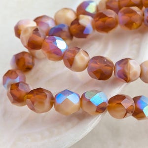 May include: Close-up of strands of faceted beads in shades of amber, peach, and cream. The beads have an iridescent sheen, reflecting hues of blue and purple. The beads are likely for jewelry making.