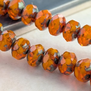 May include: Two parallel strands of faceted, translucent orange glass beads. Each bead features a round, flattened shape with a polished finish and subtle metallic gray inclusions embedded within the glass, creating a reflective, multi-faceted surface.