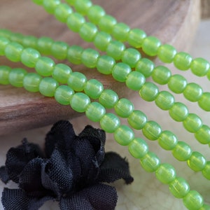 May include: A strand of lime green glass beads, with a slight iridescent sheen, is displayed on a wooden surface. The beads are round and have a smooth, polished finish.