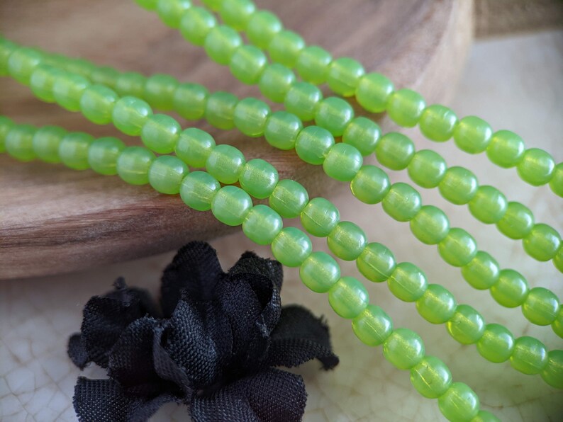 May include: A strand of lime green glass beads, with a slight iridescent sheen, is displayed on a wooden surface. The beads are round and have a smooth, polished finish.