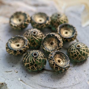 May include: A collection of artisan beads with a unique design. Each bead features a gold-colored interior and a dark green exterior with a gold-colored, geometric pattern. The beads are round with a hole for stringing, perfect for jewelry making.