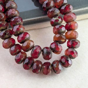 May include: A strand of faceted, round beads in deep shades of burgundy and dark red. The beads feature a translucent quality with subtle, darker inclusions and a polished, reflective surface, arranged in a curved, multi-row formation.