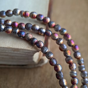 May include: Close-up of three strands of iridescent beads. The beads are a mix of gray, purple, and gold, with a textured surface. The beads are draped over the edge of a vintage book, with a wooden background.