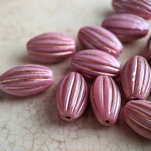 May include: A collection of pink and gold oval-shaped beads with a ribbed texture. The beads are arranged in a pile on a light brown surface.