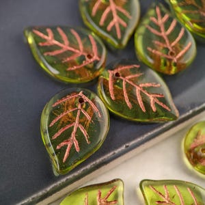 May include: A collection of green glass leaf-shaped beads with a copper-colored vein design. The beads have a hole in the top for stringing.