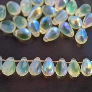 May include: A strand of iridescent green glass teardrop beads. The beads are strung on a thin wire and are arranged in a row. The beads are all the same size and shape, and they have a pearlescent finish.