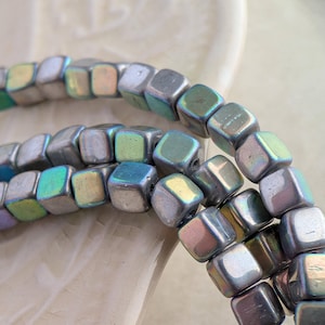 May include: A strand of small, cube-shaped beads with a metallic, iridescent finish. The beads display shifting hues of silver, green, blue, and soft purple, reflecting light across their smooth, rounded edges and uniform, geometric square shapes.