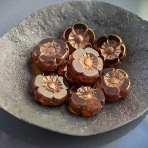 May include: A collection of eight copper-colored glass flower-shaped beads. The beads are arranged in a small metal bowl.