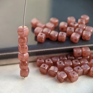 May include: A strand of small, square, pink beads with a metallic sheen. The beads are strung on a wire and are arranged in a vertical stack. There are also loose beads scattered around the stack.