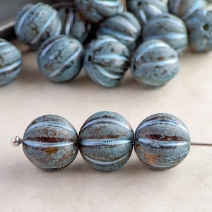 May include: A collection of teal and brown round beads with white stripes. Three beads are strung on a silver needle, while others are scattered in the background. The beads have a textured, weathered appearance, suggesting a vintage or handcrafted style.