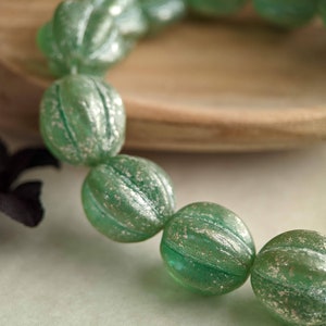 May include: A close-up of a strand of green glass beads. The beads are round with a ribbed texture and a silver-colored shimmer. The beads are strung together, and the background includes a wooden bowl and a black fabric.