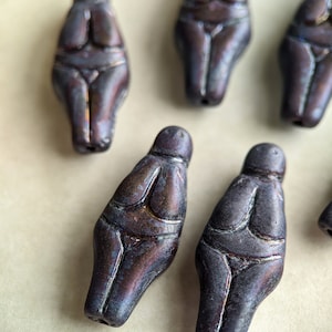 May include: Close-up of several small, dark brown Venus figurines. The figurines have a rounded shape with carved details representing the body. The color has hints of purple and blue. The figurines are arranged on a light-colored surface.