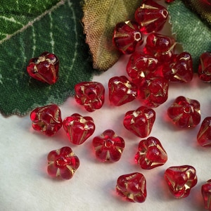 May include: A collection of red glass beads with gold accents. The beads are shaped like flowers with five petals. The beads are scattered on a white surface.