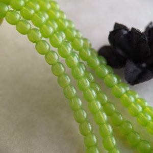 May include: A strand of green glass beads, with a slight shimmer, is shown in a close-up image. The beads are round and have a smooth, polished finish.