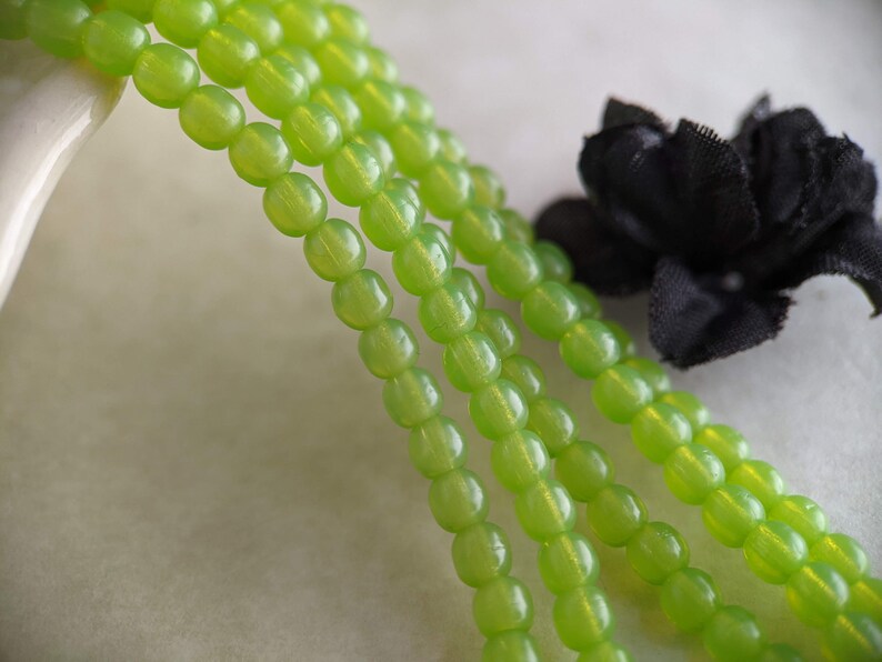 May include: A strand of green glass beads, with a slight shimmer, is shown in a close-up image. The beads are round and have a smooth, polished finish.