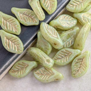 May include: A collection of pale green leaf-shaped beads with gold-colored veining. The beads are scattered on a dark gray surface, showcasing their delicate design and texture. These beads are ideal for jewelry making and craft projects.
