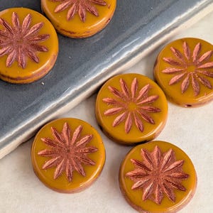 May include: Close-up of several round, mustard-yellow beads with a raised, reddish-brown floral design. The beads are evenly spaced and appear to be made of glass or a similar material. The floral design is a starburst pattern.