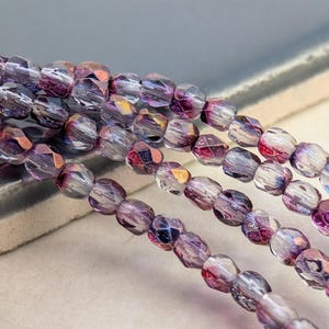 May include: Close-up of several strands of faceted beads in shades of purple, pink, and clear. The beads have a geometric shape and reflect light, creating a sparkling effect. The beads are strung together, suggesting they are for jewelry making.