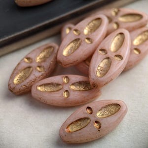 May include: A group of oval-shaped glass beads with a pink base and gold accents. The beads have a raised design of four gold circles in the center.