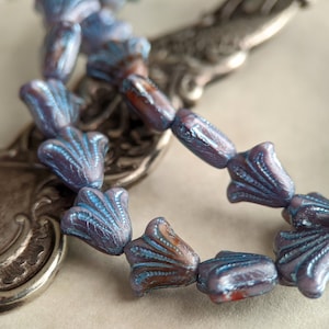 May include: A strand of iridescent blue glass beads with a flower-like design. The beads are strung on a silver chain and are lying on a silver surface.