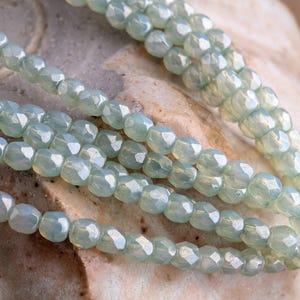May include: Close-up of several strands of light blue faceted beads. The beads have a pearlescent sheen and are strung together, creating a delicate and elegant appearance. The background is a neutral, textured surface.