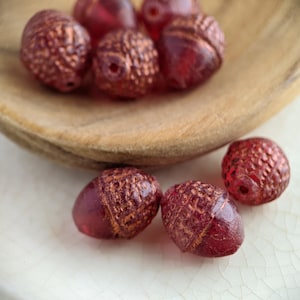 May include: Close-up of several red-toned glass beads with a textured surface, resembling acorns. Some beads are in a wooden bowl, others are scattered on a white surface. The beads have a hole for stringing.