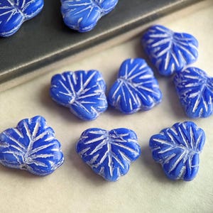 May include: A collection of eight blue glass beads in the shape of leaves. The beads have a white, textured design.