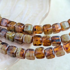 May include: A strand of square-shaped glass beads featuring a marbled pattern in shades of amber, brown, and cream. The beads are arranged in multiple rows, showcasing a smooth, polished finish with translucent and opaque color variations.