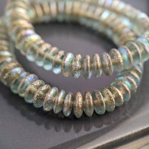 May include: Strands of iridescent, disc-shaped beads. The beads have a light blue and gold color with a textured, metallic finish. The beads are strung together, forming a circular shape, suitable for jewelry making.