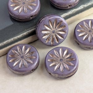 May include: Close-up of several round, lavender-colored beads. Each bead features a raised, silver-colored floral design in the center. The beads have a slightly distressed appearance, with visible texture and subtle variations in color.