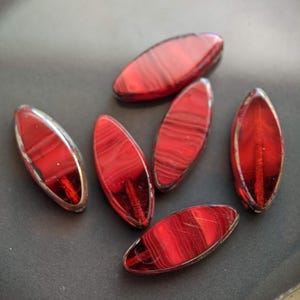 May include: Six red glass beads with a striped pattern. The beads are oval shaped and have a glossy finish.