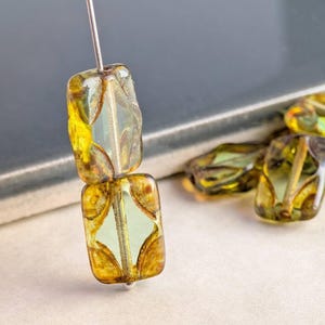 May include: Two rectangular, transparent beads with a yellow and brown design are strung on a silver pin. Several similar beads are scattered in the background. The beads have a unique, artistic appearance, suitable for jewelry making.