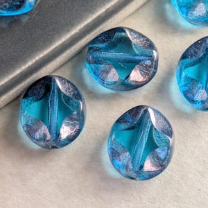May include: Close-up of several blue glass beads. Each bead has a star-shaped design in the center and a faceted, oval shape. The beads are a vibrant turquoise color with silver accents, and are scattered on a light surface.