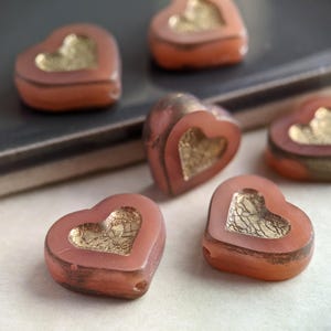 May include: Five pink heart-shaped glass beads with gold foil accents. The beads are arranged on a dark surface.