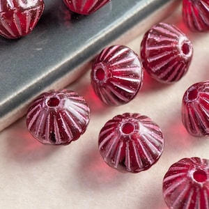 May include: Close-up of several faceted, dark red glass beads. Each bead has a central hole and is decorated with vertical, light pink lines. The beads are scattered on a light surface, with a gray, rectangular object in the background.