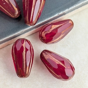 May include: Close-up of several faceted, teardrop-shaped beads. The beads are a deep red color with a metallic gold-colored stripe running down the center. The beads are arranged on a light-colored surface.