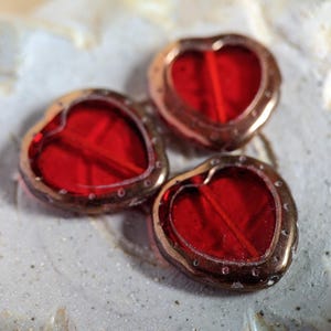 May include: Three heart-shaped beads with a vibrant red center and a bronze-colored metal frame. The beads are arranged on a textured, off-white surface, creating a romantic and handcrafted feel. These beads are ideal for jewelry making.