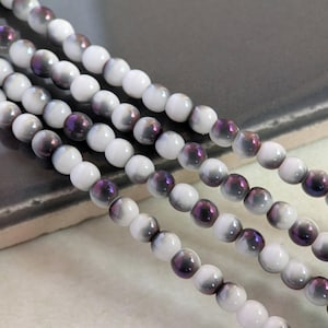 May include: Strands of white and iridescent purple beads. The beads are round and uniform in size, strung together to create a necklace or bracelet. The beads have a pearlescent sheen, reflecting light.