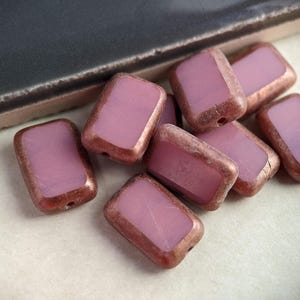 May include: A group of eight rectangular glass beads with a pink, translucent color and a copper-toned metallic rim.