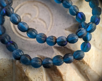 May include: A strand of blue glass beads with a textured surface. The beads are oval shaped and have a slight iridescent sheen.