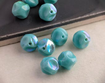 May include: A collection of iridescent turquoise beads with faceted surfaces. Each bead has a small hole for stringing. The beads are scattered on a light-colored surface, with some resting near a dark, rectangular object.