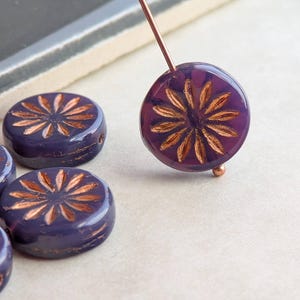 May include: Close-up of several dark purple, flat, round beads with a copper-colored floral design. One bead is strung on a copper wire. The beads are likely for jewelry making.