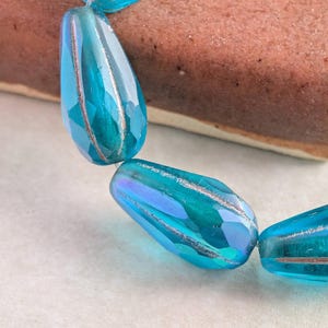 May include: Close-up of a strand of faceted, teardrop-shaped beads. The beads are a vibrant turquoise color with silver-colored stripes. The background includes a brown, rectangular object and a white surface.