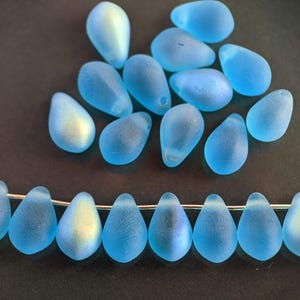May include: A strand of light blue glass beads in a teardrop shape. The beads have a frosted finish and a subtle iridescent shimmer.
