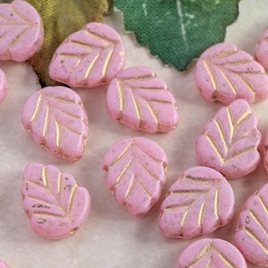 May include: A collection of small, pink, leaf-shaped beads scattered across the frame. Each bead features a textured surface with recessed gold-colored veins that highlight the leaf pattern. The beads are opaque and have a matte finish.