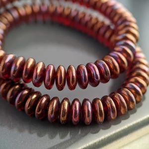 May include: Close-up of two strands of iridescent, reddish-brown beads. The beads are small, flat, and disc-shaped, with a metallic sheen. The beads are arranged in a circular pattern, suggesting they are part of a necklace or bracelet.