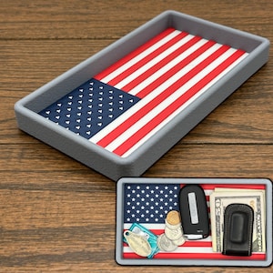 May include: A rectangular gray tray with an American flag design on the inside. The flag features red and white stripes and a blue field with white stars. A smaller tray with items like keys, money, and a wallet is also visible.
