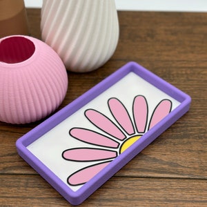 May include: A purple rectangular tray with a white background and a pink daisy design. The daisy has a yellow center.