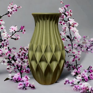 May include: A green ceramic vase with a geometric pattern. The vase is sitting on a white surface with pink flowers in the background.